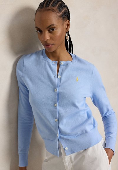 Frau mit geflochtenem Haar, die eine hellblaue, geknöpfte Strickjacke und weiße Hose trägt und vor einer einfarbigen beige Wand steht.