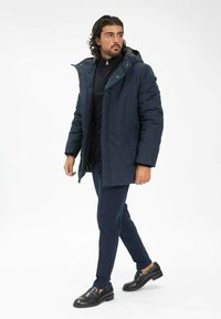 Doudoune marine avec capuche, fermeture éclair à l'avant et poches latérales ; associée à un pantalon slim bleu foncé et des chaussures en cuir noires.