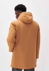 Parka marrón con capucha, de textura suave y mate; mangas ajustadas; detalles en la costura trasera; y una silueta ligeramente más larga.