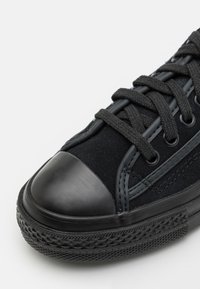 Svart sneaker med gummikappa vid tån, texturerad överdel och platta snören. Har detaljer med sömnad och en mönstrad gummisula.