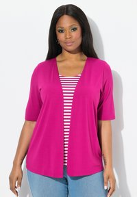 Cardigan fucsia con abertura frontal y mangas cortas, superpuesto sobre una camiseta de rayas blanca y roja. Llevado con jeans de mezclilla azul claro.