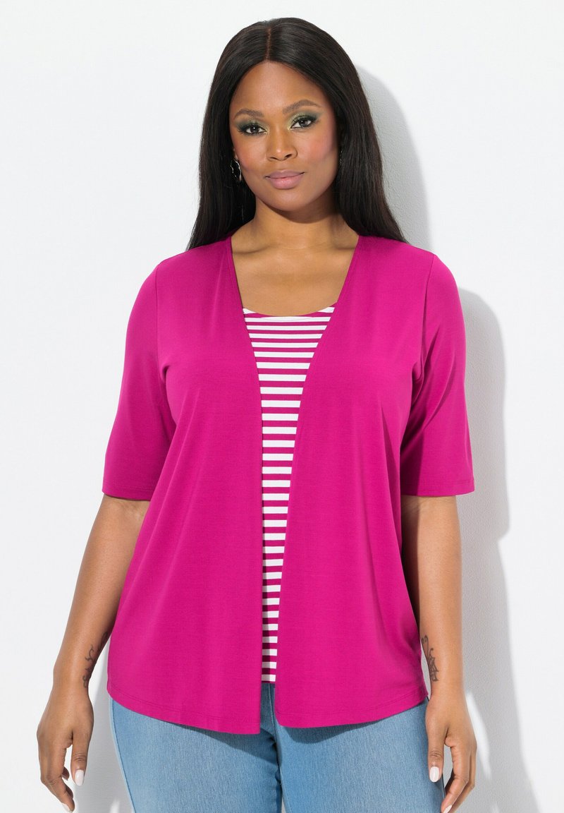 Cardigan fucsia con abertura frontal y mangas cortas, superpuesto sobre una camiseta de rayas blanca y roja. Llevado con jeans de mezclilla azul claro.