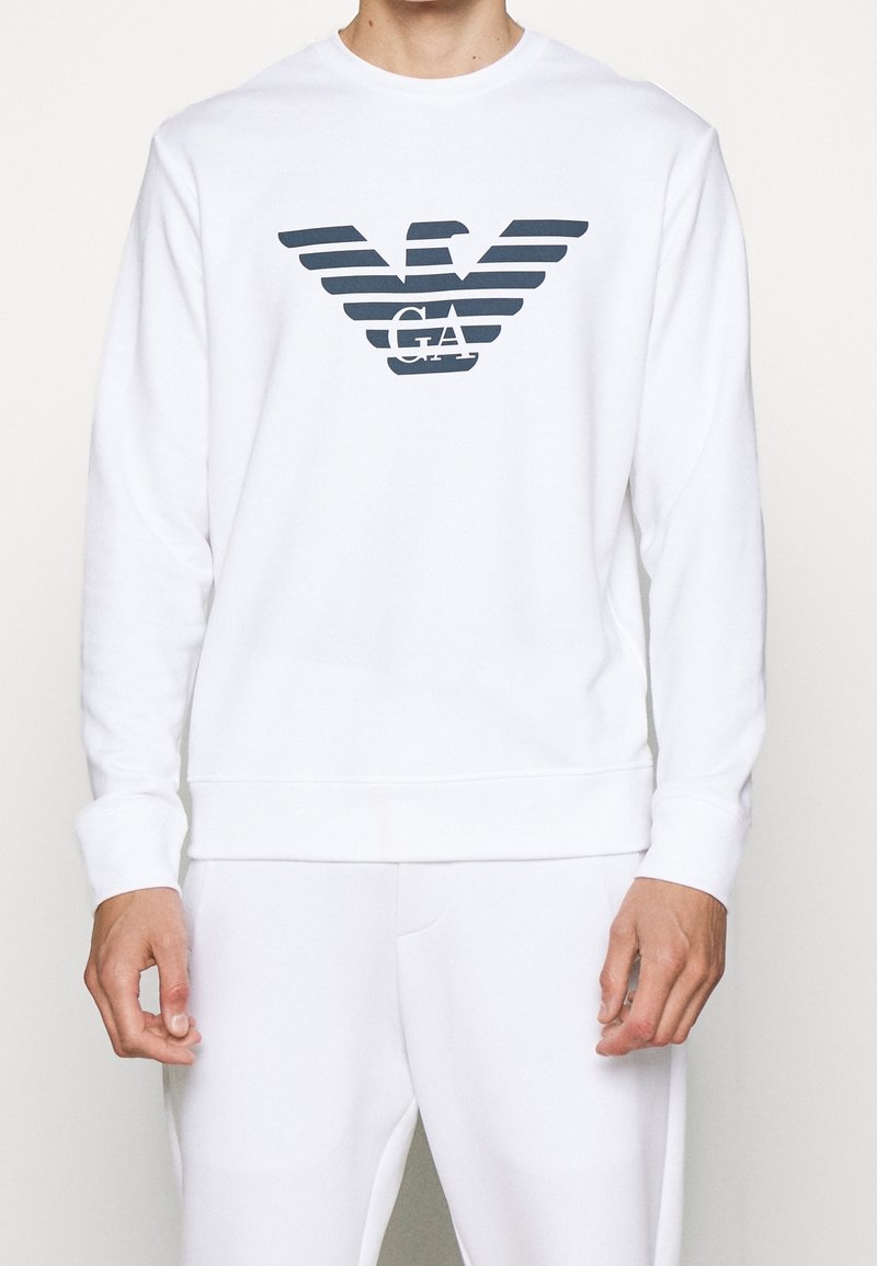 Vit sweatshirt med en mörkblå randig örnlogga och "GA" under, med ribbade muddar och nederkant, tillverkad av mjukt tyg.