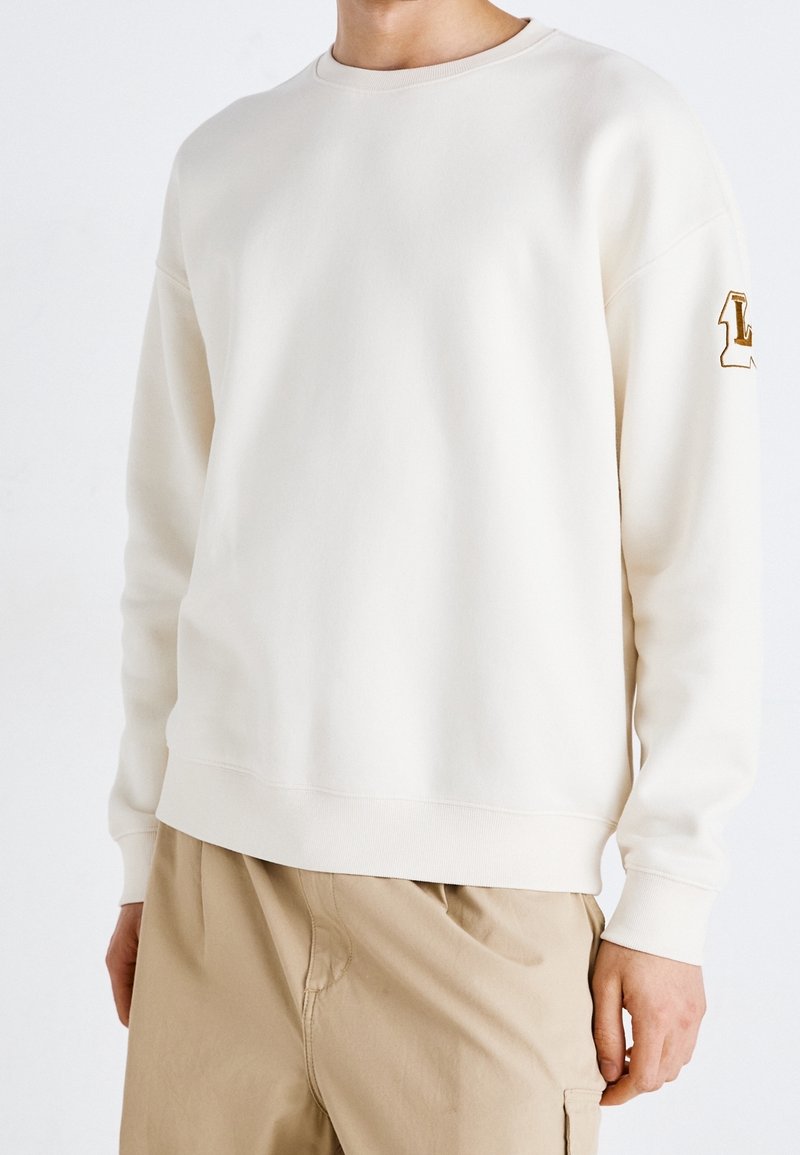 Homme portant un sweat-shirt blanc uni à manches longues avec des poignets côtelés et un pantalon beige, debout devant un fond blanc.