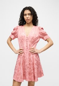 Γυναίκα με κατσαρά μαλλιά, που φοράει ροζ φόρεμα με λουλούδια, κουμπωτό και με φουσκωτά μανίκια, στέκεται με τα χέρια στη μέση μπροστά από ένα απλό φόντο.