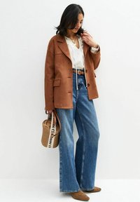 Bruine suède blazer over een witte top, gedragen met hoog-gewicht denim flared jeans en bruine enkelhoge laarzen. Accessoires zijn onder andere een geweven tas.