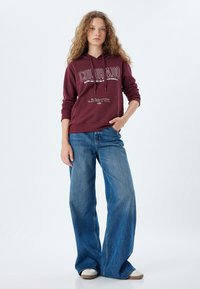 Bordowy sweatshirt z kapturem z napisem "COLORADO" w kolorze białym, w połączeniu z jasnoniebieskimi długimi dżinsami i białymi trampkami.