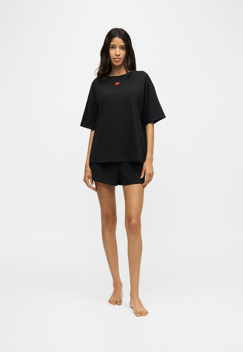 Femme debout pieds nus, portant un t-shirt noir ample et un short noir sur un fond blanc uni.