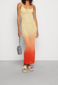 Strick-Maxi-Kleid mit Gelb-Orange-Verlauf, ausgestattet mit dünnen Trägern und einer figurbetonten Form. Kombiniert mit beigen Plateausandalen und einer grauen Tasche.