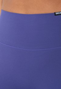 Violetti korkeavyötäröiset legginsit, jotka on valmistettu sileästä kankaasta. Niissä on ommeltu vyötärönauha ja pieni tuotemerkki vasemmassa yläkulmassa.
