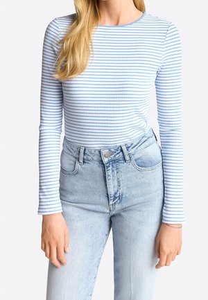 Femme portant un t-shirt côtelé à manches longues rayé bleu clair et blanc, rentré dans un jean taille haute délavé clair.