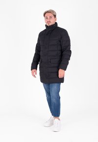 Giubbotto imbottito nero con colletto alto, forma boxy, cuciture orizzontali e due tasche frontali. Indossato con jeans blu e sneaker bianche.