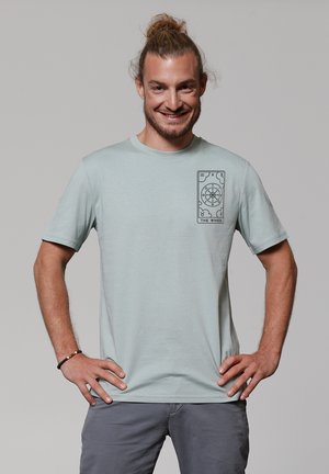 Helles blaues T-Shirt mit kurzen Ärmeln aus weichem Material, das auf der linken Brust ein schwarzes Grafikdesign zeigt, das ein Rad und Symbole darstellt.