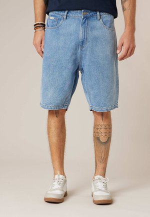 Homme portant un short en jean bleu clair, des baskets blanches, une chemise noire, avec des tatouages sur les jambes et des bracelets au poignet gauche, debout sur un fond uni.