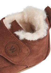 Botă din suede maro, cu o căptușeală moale din blană de miel de culoare crem și un patch logo circular. Dispune de un închidere cu cârlige și bucle pentru o purtare ușoară.