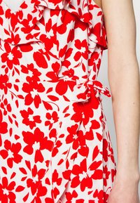 Weißes Kleid aus Stoff mit einem roten Blumenmuster, versehen mit Rüschen entlang des Ausschnitts und einer gebundenen Taillenpartie zur Formgebung.