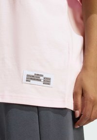 Roza pamučna majica s kratkim rukavima ima bijelu oznaku na dnu, na kojoj se nalaze crni natpisi uključujući "ADIDAS" i razne motivacijske riječi.