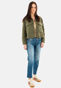 Olijfgroene cropped jas met knoopsluiting en twee voorzakken, gecombineerd met lichtblauwe spijkerbroek en beige sneakers met bordeauxrode strepen.