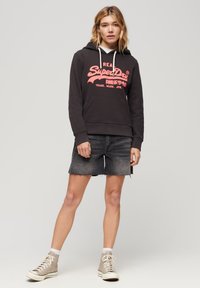 Felpa nera con cappuccio e tasca anteriore tipo kangaroo, caratterizzata da un grande logo rosa. Abbinata a pantaloni corti in denim grigio scuro e sneakers.