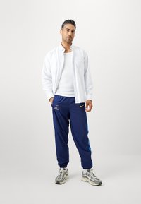 Weiß gestreiftes Hemd über einem weißen Top, kombiniert mit blauen Nike Jogginghosen mit seitlichen Akzenten und einem Logo, ergänzt durch graue Sneakers.