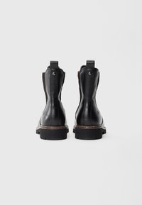 Une paire de bottines noires en cuir avec des panneaux élastiques latéraux marron et de épaisses semelles en caoutchouc, vues de l'arrière sur un fond uni.