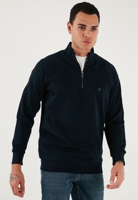 Sötétkék, magas galléros pulóver, negyed cipzárral és bordázott mandzsettákkal. Puha anyagból készült, egyszerű logó részlettel a mellkasán.