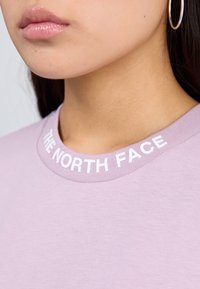Camisa de lavanda clara con cuello acanalado que presenta el texto "THE NORTH FACE" en letras blancas en negrita. Textura de tela suave, vista en primer plano.