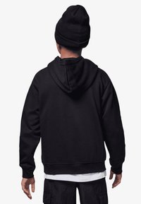 Felpa nera con cappuccio e tasca a marsupio, tessuto spesso, vestibilità comoda e polsini a coste. Indossata con un berretto nero abbinato.