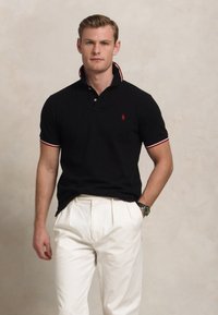 Camisa polo preta com detalhes em vermelho e branco, mangas curtas e colarinho. Combinada com calças brancas. Detalhe de marcação mínima na camisa.