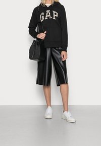 Felpa nera con zip e logo "GAP" in stampa leopardata, abbinata a pantaloni corti in ecopelle nera lucida e scarpe da ginnastica bianche, mentre tiene una borsa nera con texture.