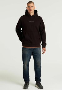 Felpa nera con cappuccio e una tasca frontale, con un logo sul petto. Indossata sopra una maglietta arancione, con jeans in denim blu e sneakers grigie.