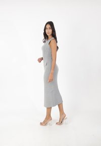 Graues, geripptes Strick-Midikleid mit ärmellosem Design, seitlichem Schlitz und Fronttaschendetails. Getragen mit klaren, hochhackigen Sandalen.