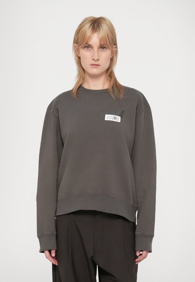 Persona rubia vistiendo un sudadera gris oscuro con un pequeño diseño de etiqueta blanca en el pecho y pantalones marrones oscuros, de pie contra un fondo liso.