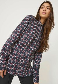 Langärmlige Bluse in Marineblau mit einem geometrischen Muster in Rot, Weiß und Creme. Verfügt über einen kragenständigen Ausschnitt und Knopfverschlüsse.