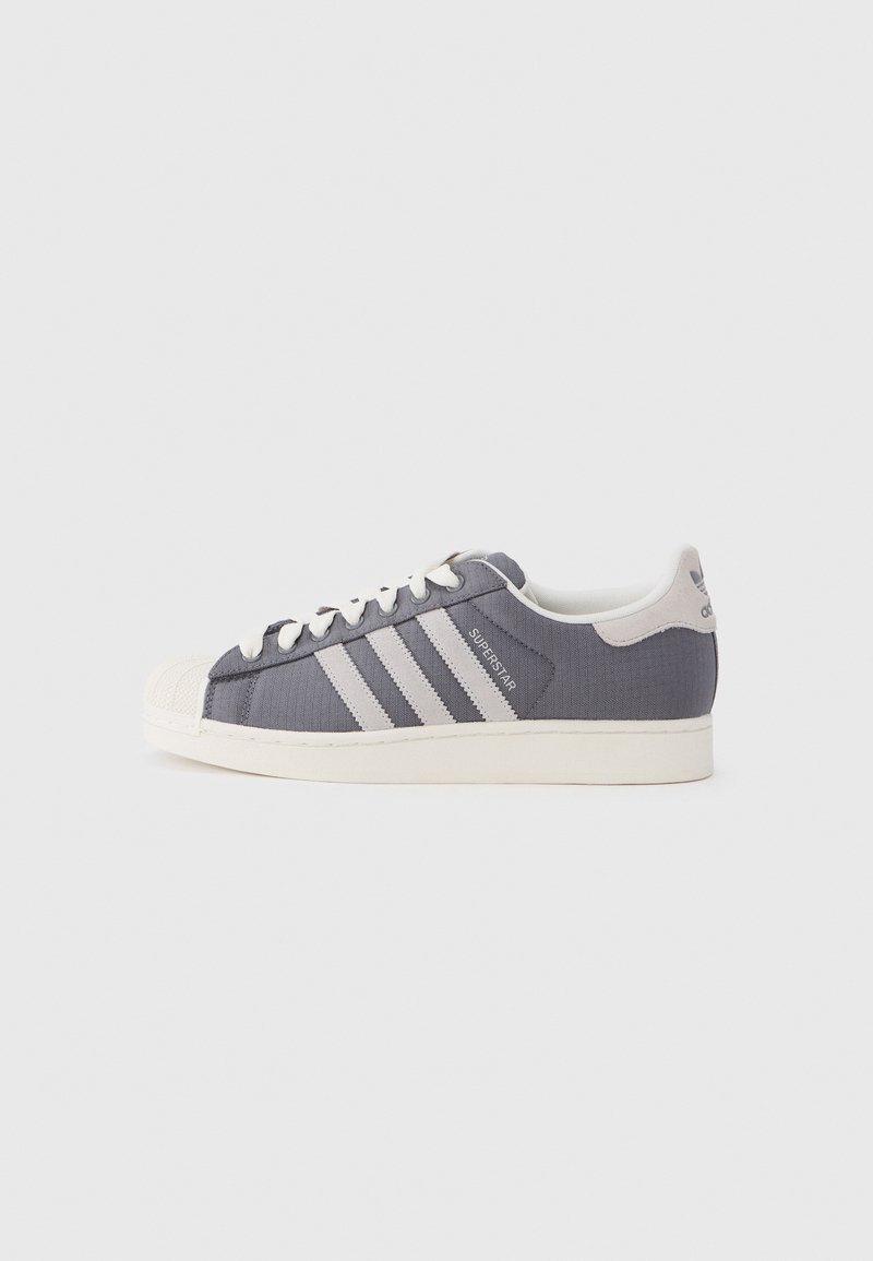Des baskets en toile grise avec trois bandes blanches horizontales, un embout en caoutchouc et un col rembourré. Lacets plats et accents de marque minimaux.