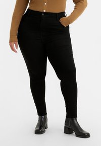 Jeans skinny negros de mezclilla con un diseño de cintura alta. Combinados con un suéter marrón de canalé y botas negras de tobillo.
