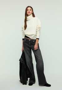 Cremfarbener Rollkragenpullover mit umgeschlagenem Kragen, kombiniert mit lockeren schwarzen Jeans und einer schwarzen Lederjacke, vor einem hellen Hintergrund.