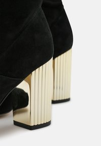 Bottes cheville en daim noir avec talons blocs dorés brillants présentant des stries verticales sur un fond blanc.