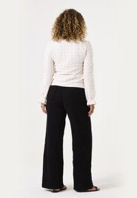 Langärmlige, weiße Bluse mit strukturierter Oberfläche im Rautenmuster und Rüschensaum, kombiniert mit schwarzen, weit geschnittenen Hosen mit hinteren Taschen.