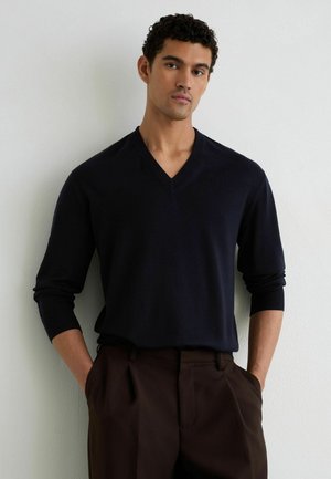 Giovane uomo con capelli ricci che indossa un maglione scuro con scollo a V e pantaloni marroni, in piedi con le mani in tasca davanti a una parete semplice.