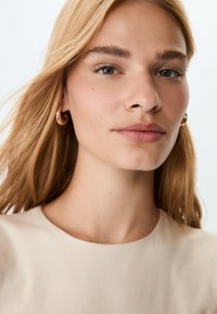 Close-up of a person with long blonde hair, wearing a beige top. Features include smooth skin, natural makeup, and gold hoop earrings.