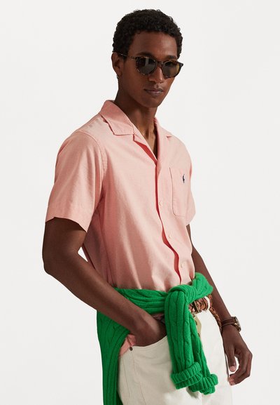 Κοντομάνικο ροζ πουκάμισο με γιακά και τσέπη στο στήθος, συνδυασμένο με ένα πράσινο πλεκτό πουλόβερ δεμένο στη μέση. Οι μπεζ σορτς και τα γυαλιά ηλίου ολοκληρώνουν την εμφάνιση.