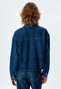 Giacca di jeans blu scuro con un design corto, polsini con bottoni e intricate ricami floreali neri lungo l'orlo.