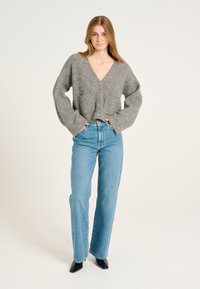 Grauer Strickcardigan mit V-Ausschnitt, Fronttaschen und fünf Knöpfen, kombiniert mit hellblauen, hochtaillierten Jeans und schwarzen spitzen Schuhen.