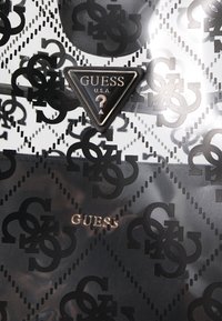 Czarno-biały dodatek o błyszczącym wykończeniu, z wzorem w diamenty i złotym logo "GUESS". Wyraźnie widoczne trójkątne metalowe logo.