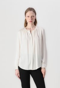 Blusa morbida a maniche lunghe in tessuto bianco avorio, con scollatura rotonda, chiusura con bottoni e sottili arricciature sulle spalle. Texture liscia.