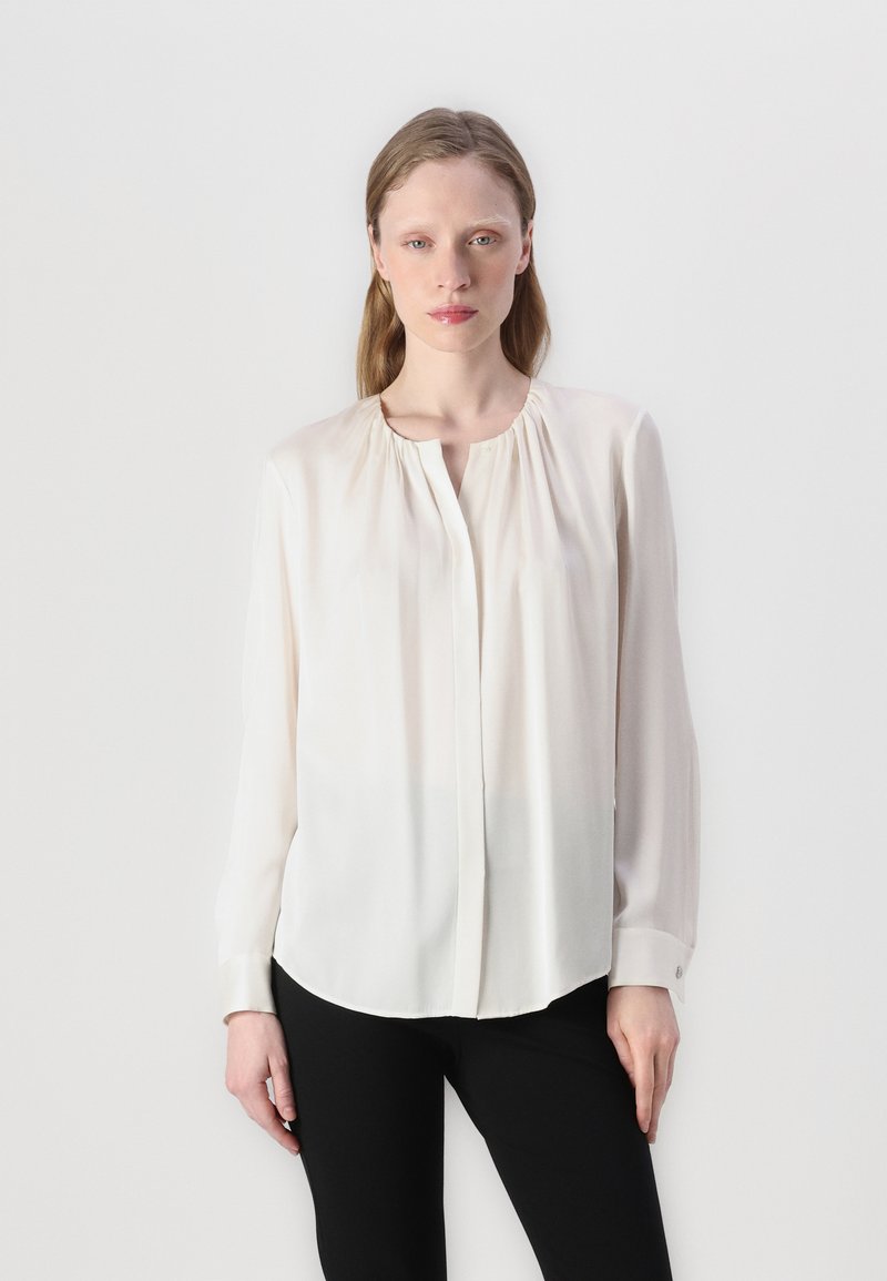 Blusa morbida a maniche lunghe in tessuto bianco avorio, con scollatura rotonda, chiusura con bottoni e sottili arricciature sulle spalle. Texture liscia.
