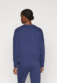 Tmavě modrá mikina s kulatým výstřihem, spadenými rameny a žebrovanými manžetami. Vyrobena z měkkého materiálu s volným střihem.