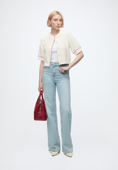Femme aux cheveux blonds courts portant un cardigan court blanc, un haut blanc, un jean bleu clair, tenant un sac à main rouge, debout sur un fond blanc.