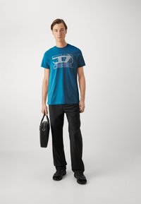 Teal bomull t-skjorte med grafisk "DIESEL" logo, kombinert med svarte bukser og svarte treningssko. Holder en svart håndveske.